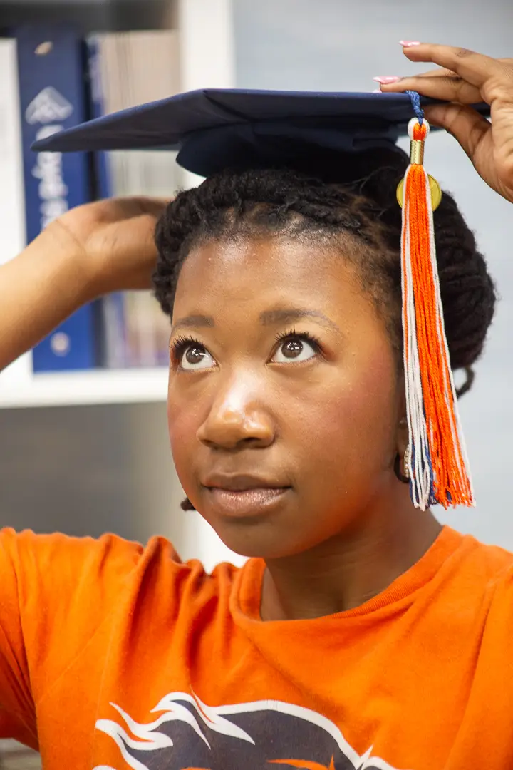 Graduate in an orange university shirt lifting her cap onto her head, photographed close and looking up.