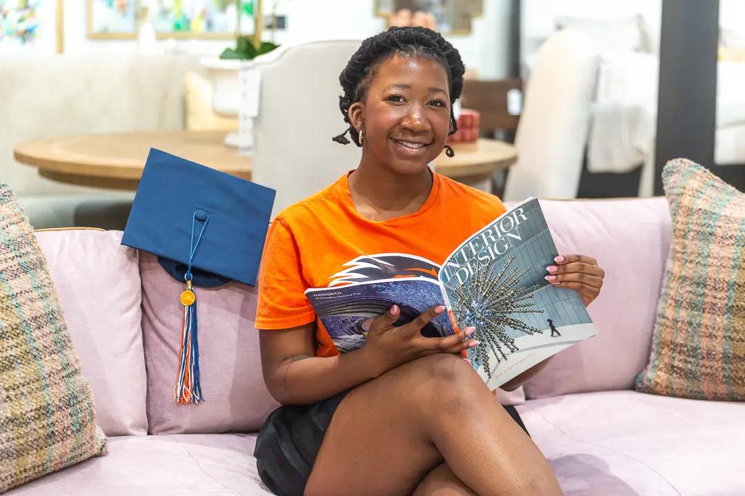 Graduate seated on a showroom sofa reading a design magazine, her cap resting beside her.