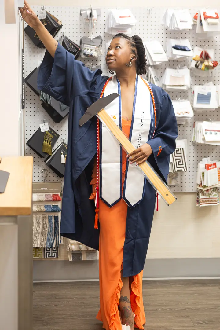 Graduate in a navy gown and orange dress reaching toward a wall of design samples, photographed by The McKinnis Edit.