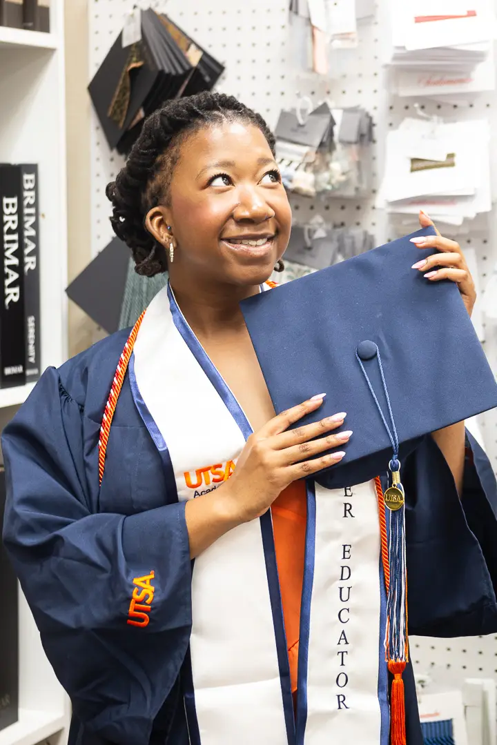 Graduate in a navy gown holding her cap up in celebration, photographed on location in a design showroom.