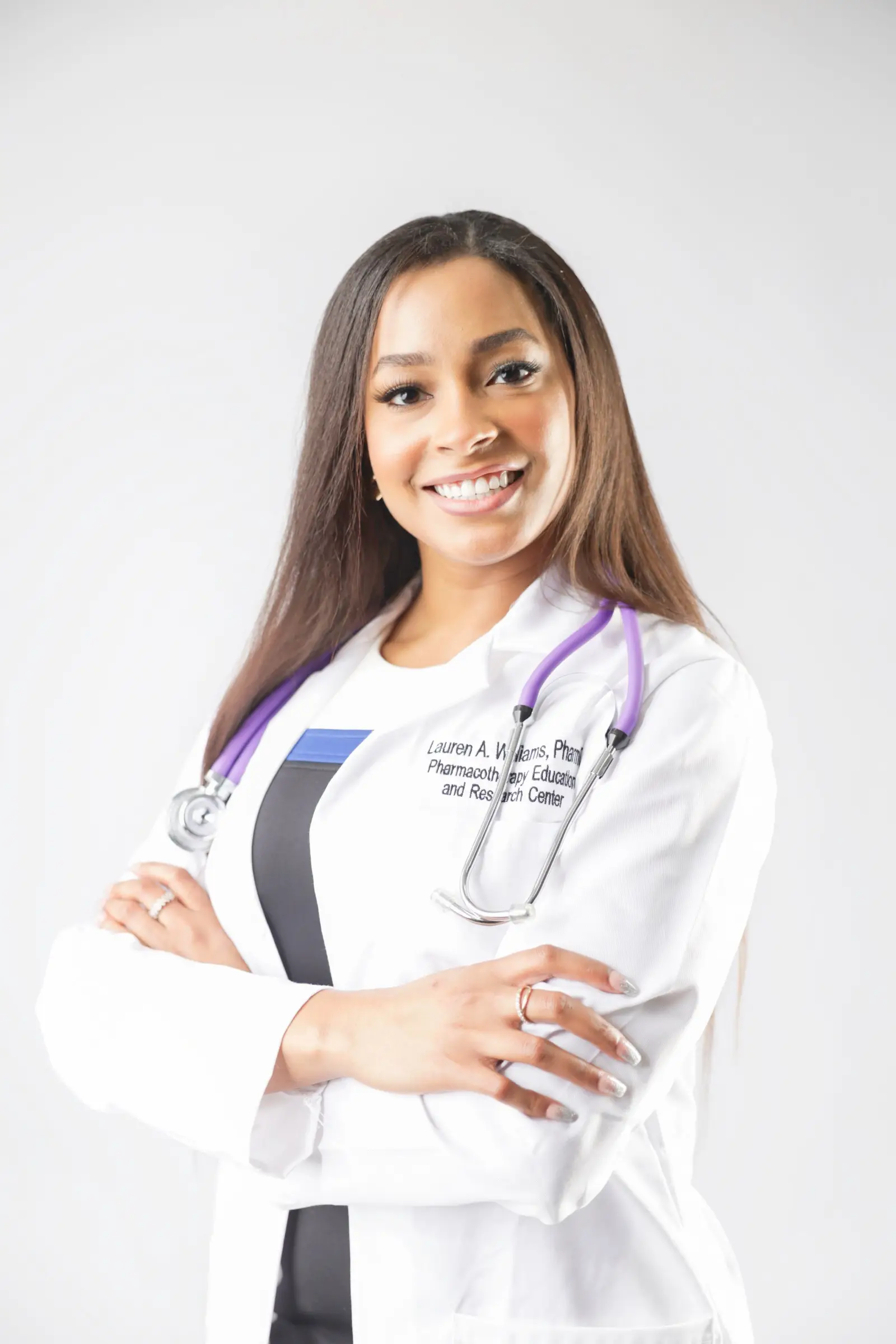Branding headshot of a pharmacist in a white coat with a stethoscope, arms crossed, smiling against a light gray backdrop.