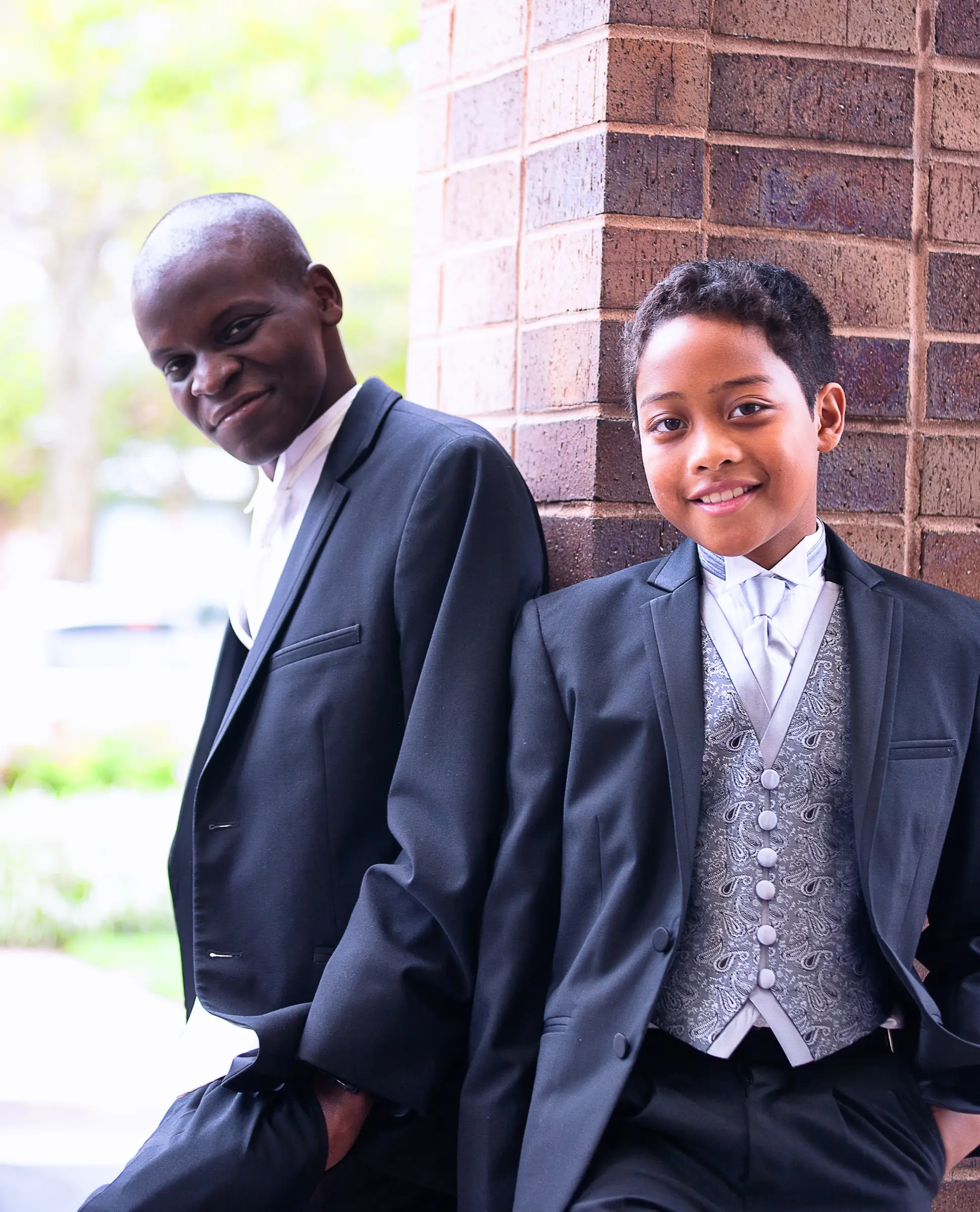 Wedding portrait of a father and son in matching formalwear, leaning against a brick column.